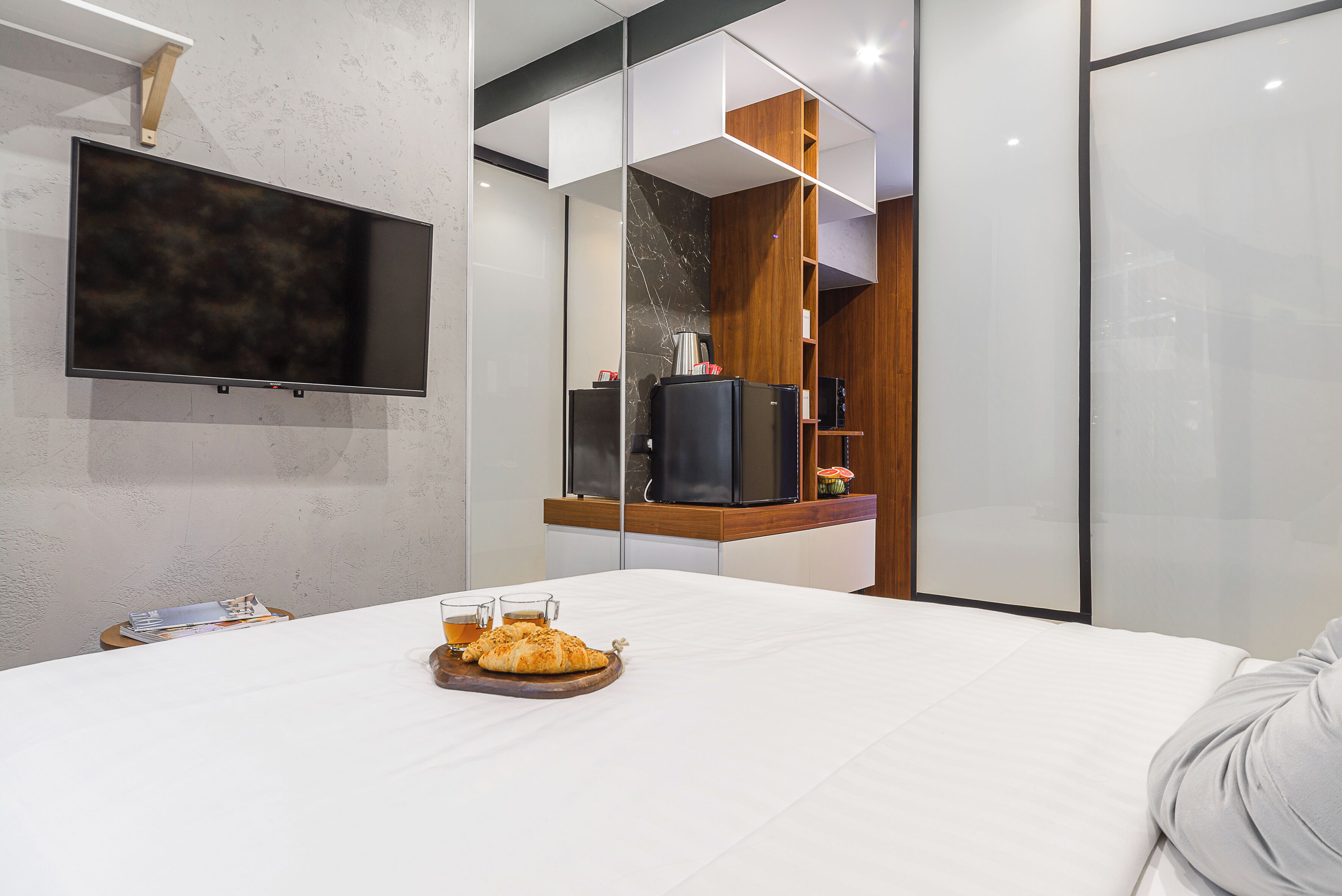 A modern hotel room with a large bed, a flat-screen TV mounted on the wall, a wooden sideboard, and a glass partition.