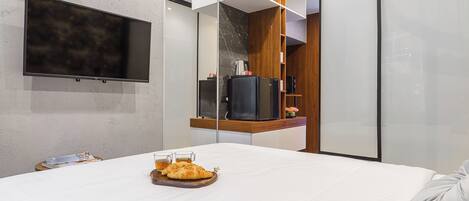 A modern hotel room with a large bed, a flat-screen TV mounted on the wall, a wooden sideboard, and a glass partition.