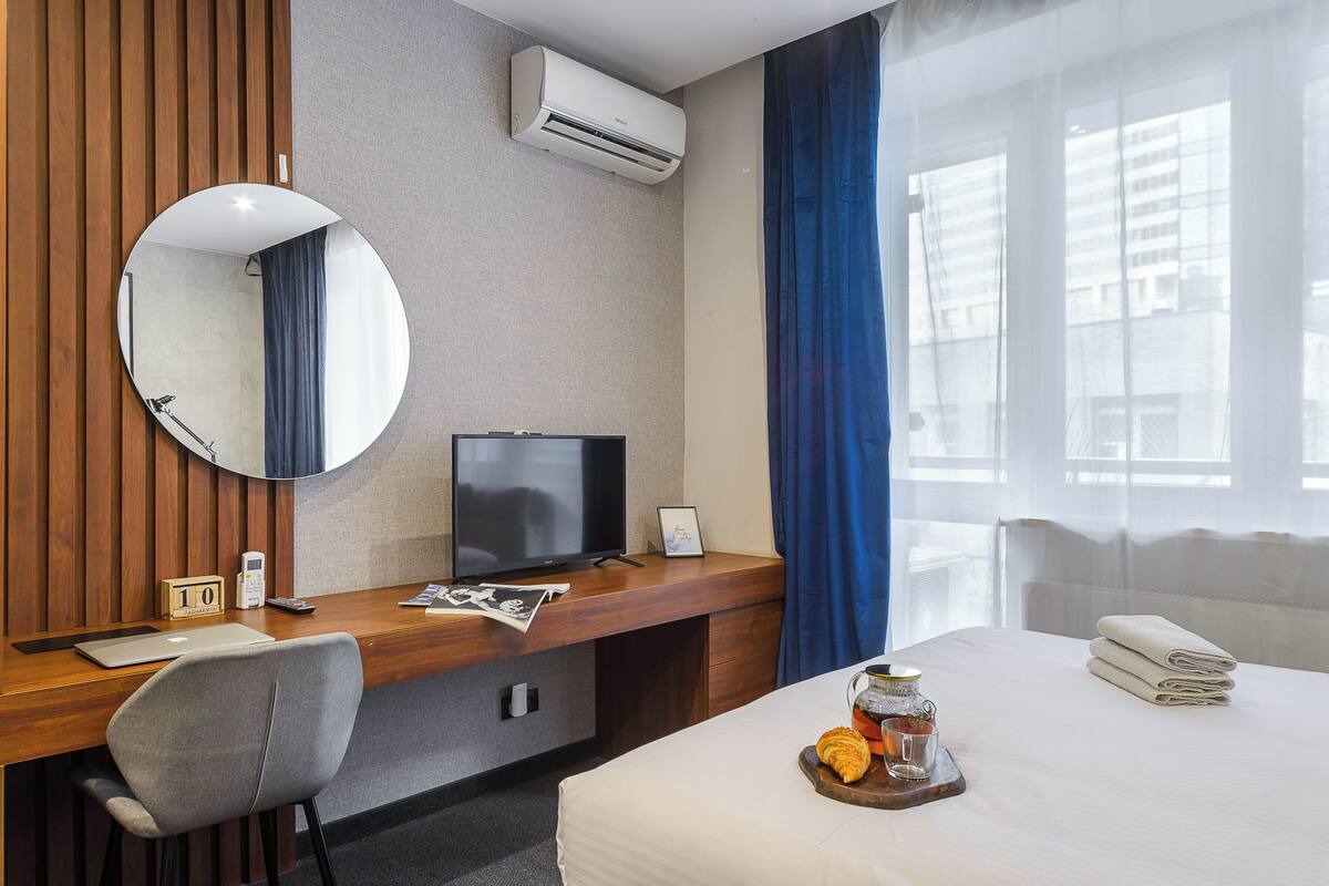 A hotel room with a wooden desk, a television, a bed with a white comforter, a round mirror, and a window with blue curtains.