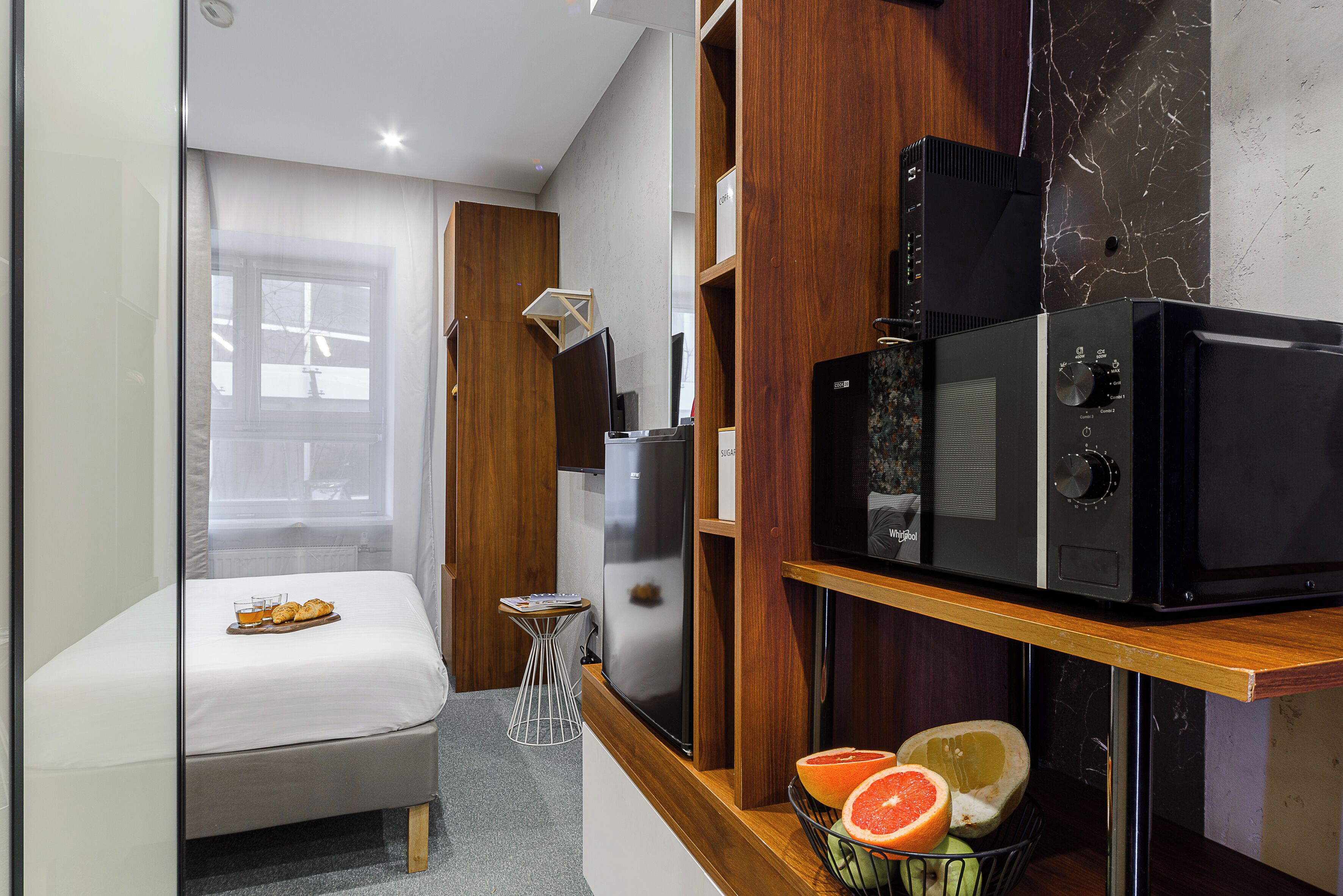 Una kitchenette compacta con microondas, un refrigerador pequeño y un estante de madera con frutas.