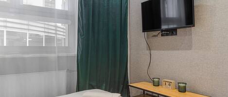 Habitación de hotel con cama, televisor montado en la pared, una mesita con un reloj y una ventana con cortinas.