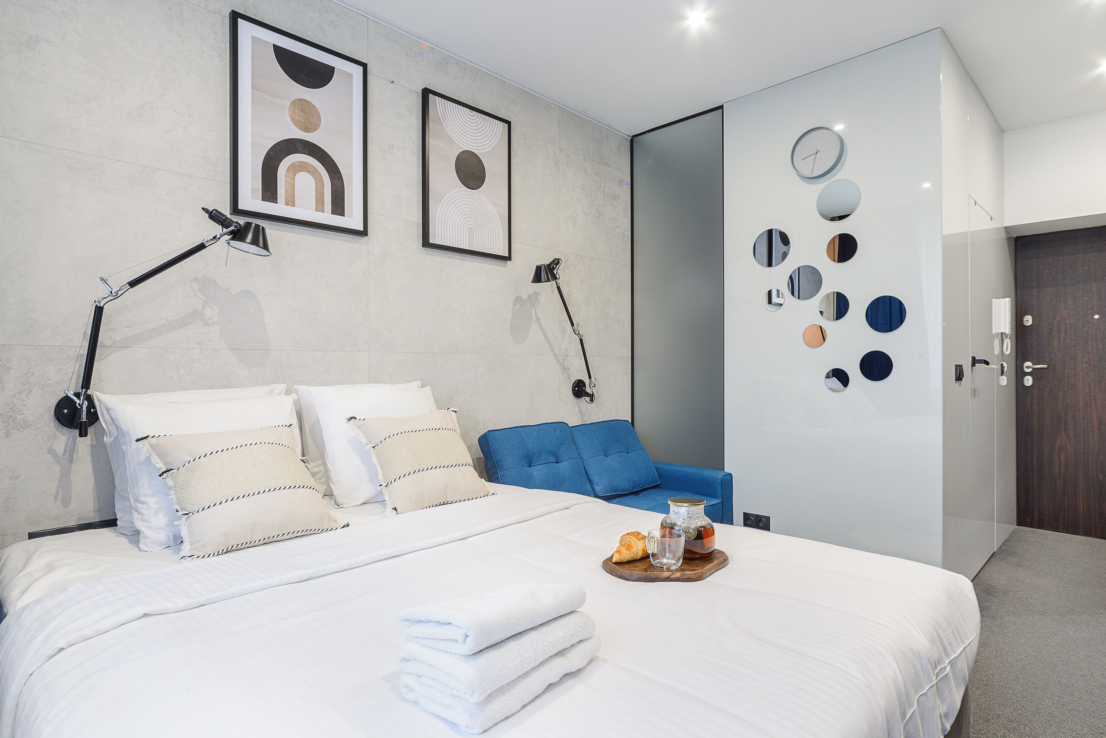 A modern hotel room with a neatly made bed, a blue sofa, a wall clock, and a tray with a croissant and a glass of water.