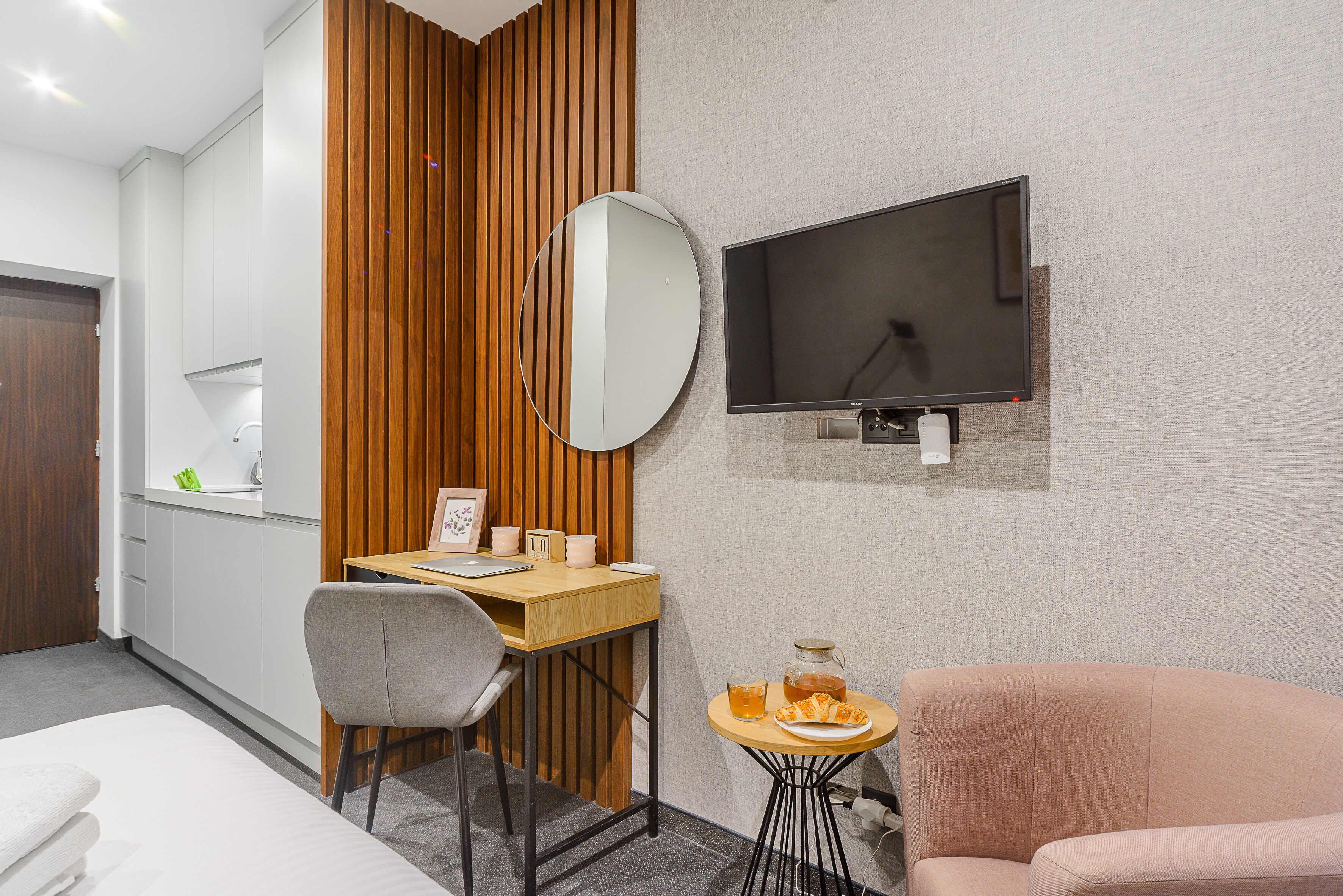 A hotel room with a bed, a desk with a chair, a small round table with a drink and snacks, a TV mounted on the wall, and a wooden panel wall.