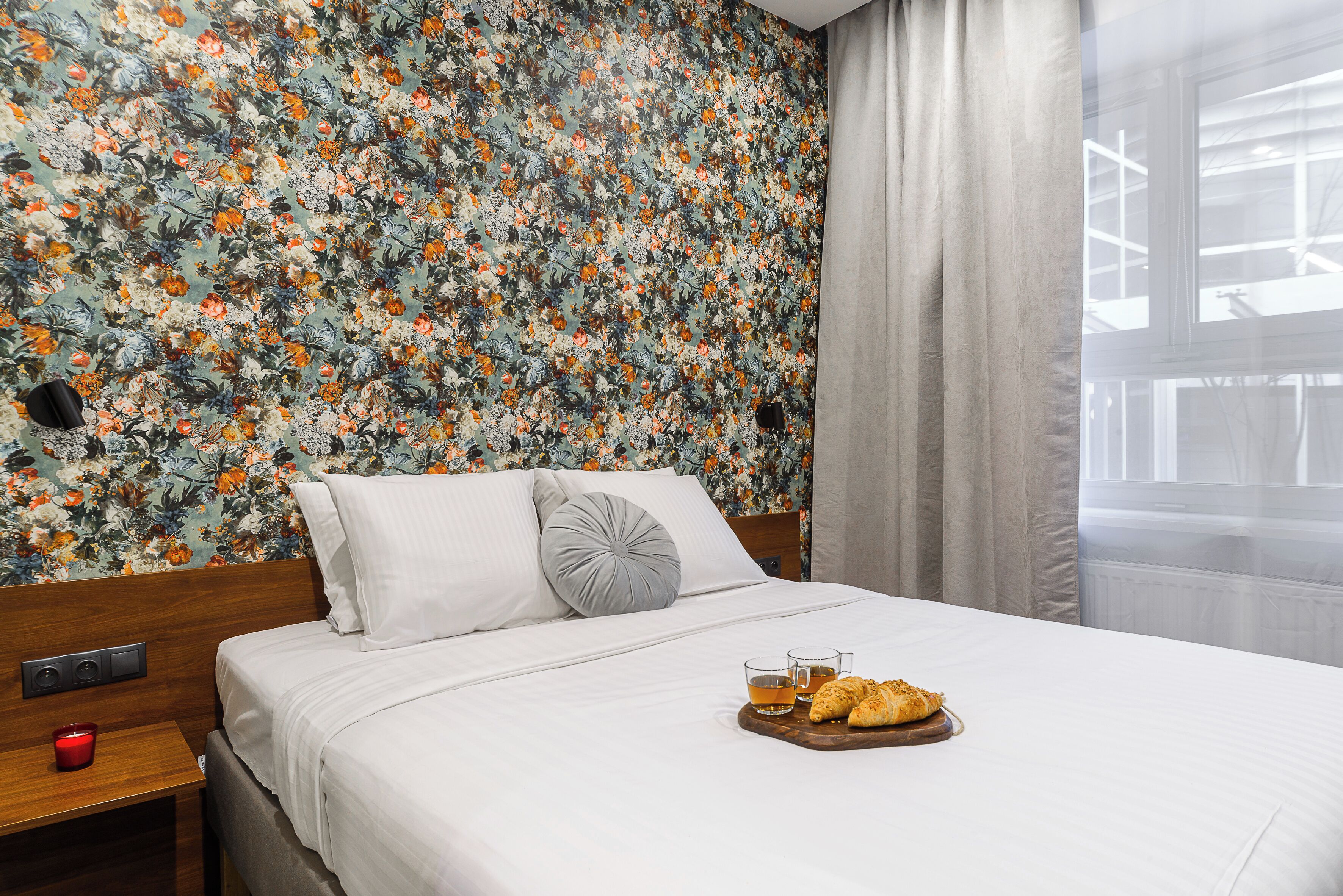 Een netjes opgemaakt bed met witte lakens, bloemenbehang, een houten nachtkastje met een rode kaars, en een dienblad met croissants en thee op het bed.