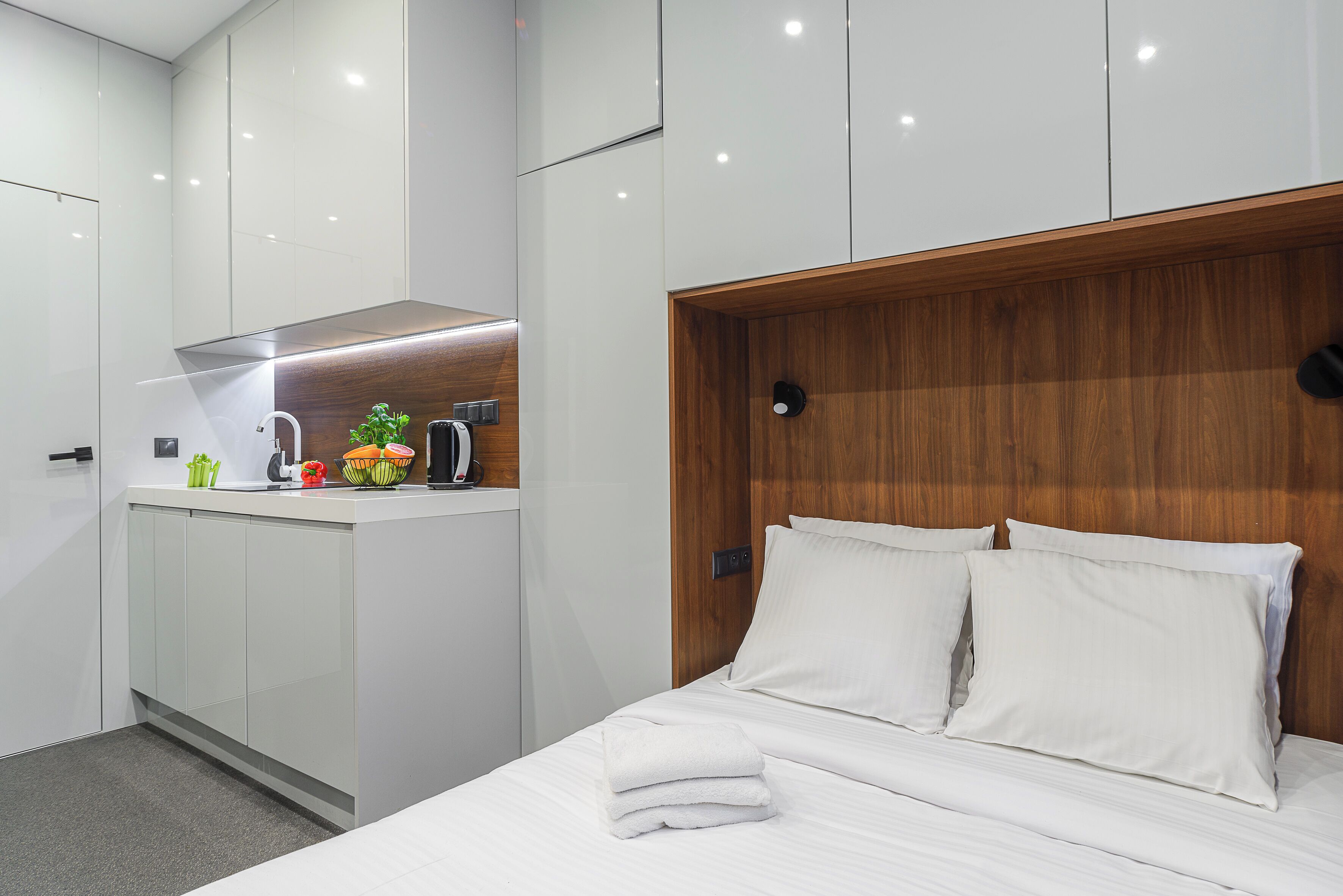 Ein modernes Hotelzimmer mit einem ordentlich bezogenen Bett, einem Holz-Kopfteil und einer kleinen Küchenzeile mit einer Mikrowelle und einer Obstschale.