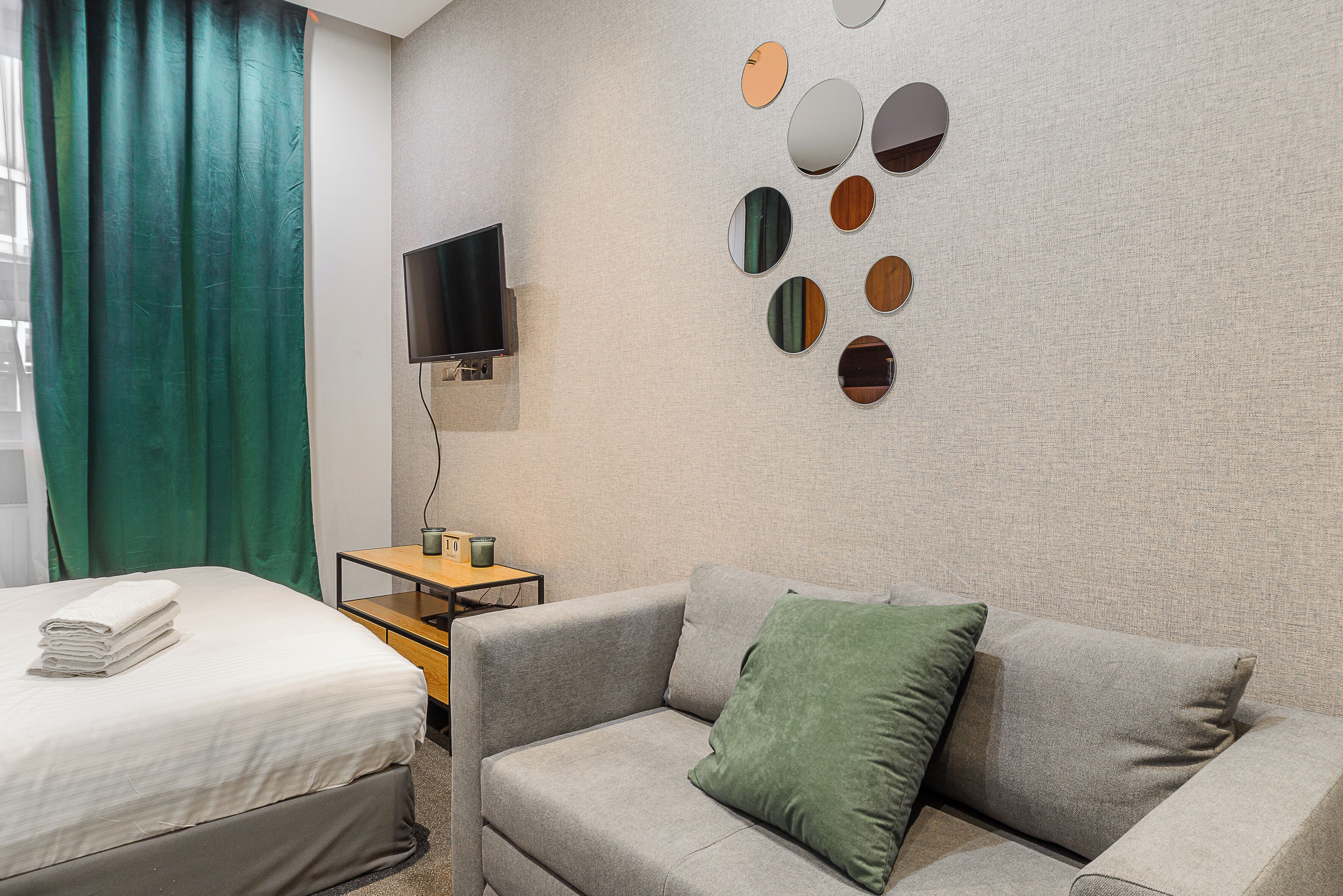 A hotel room with a bed, a grey sofa with a green pillow, a TV on the wall, and a mirror wall.