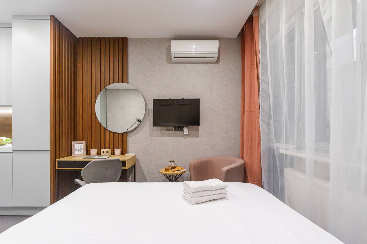 A hotel room with a bed, a desk with a chair, a round mirror, a television, and a small table with a glass and a fruit plate.