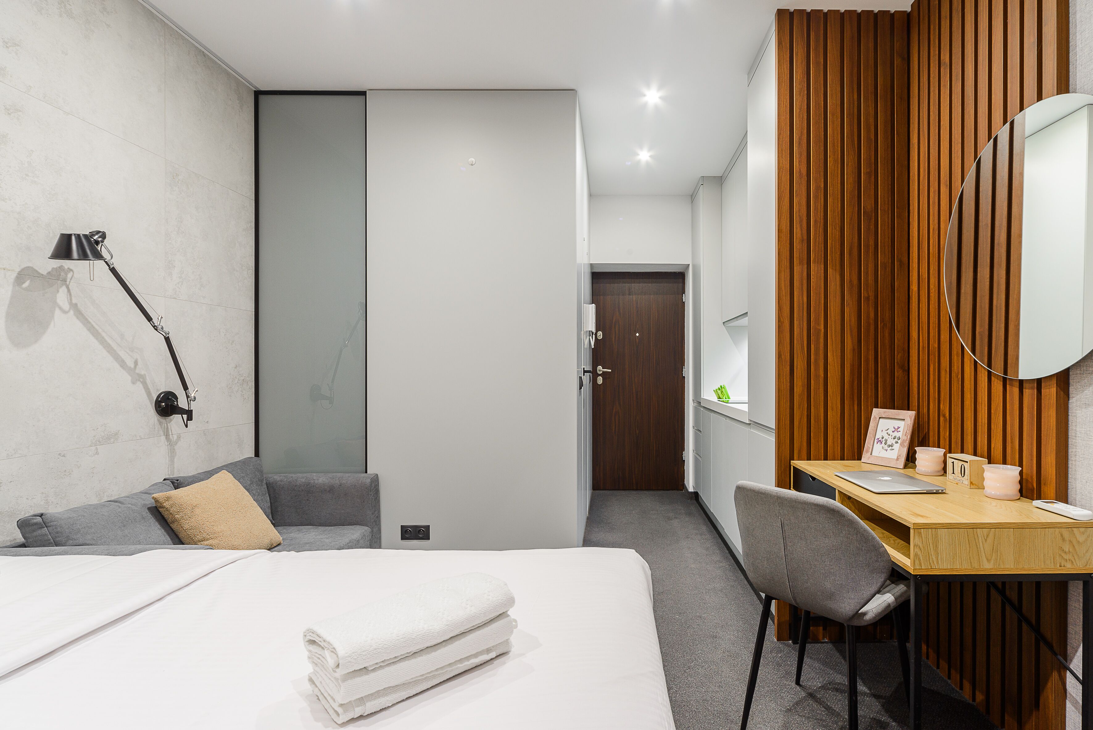 A modern hotel room with a bed, a desk, a chair, a lamp, and a wooden panel wall.