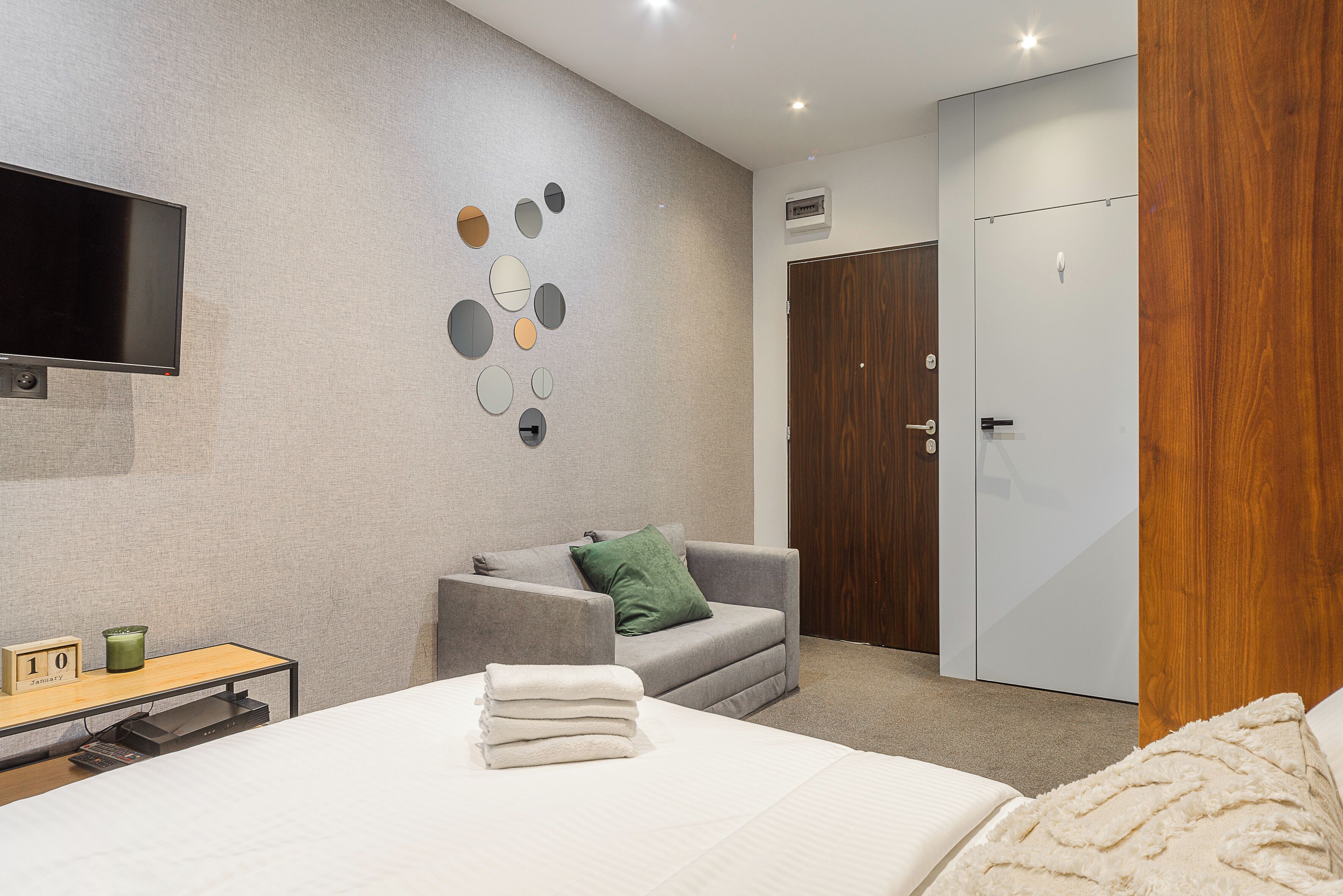A modern hotel room with a bed, a desk, a grey sofa, a TV mounted on the wall, and a wooden wardrobe.