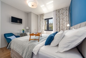 A hotel room with a bed, a TV mounted on the wall, a desk with a lamp, and a window with curtains.