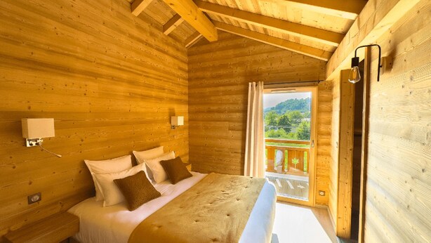 Un dormitorio de cabaña de madera con una cama, almohadas, una mesita de noche, una ventana con cortinas y vistas al verdor exterior.