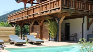 Una terraza de madera con sillones, una piscina y montañas al fondo.