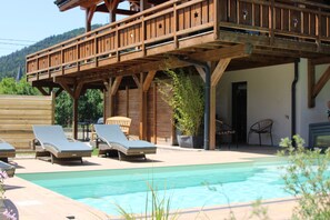 A wooden deck with lounge chairs, a pool, and a mountain backdrop. - Chalet de L'Oseraie – Swimming pool, Spa, Sauna – Vosges (LE SAULCY)