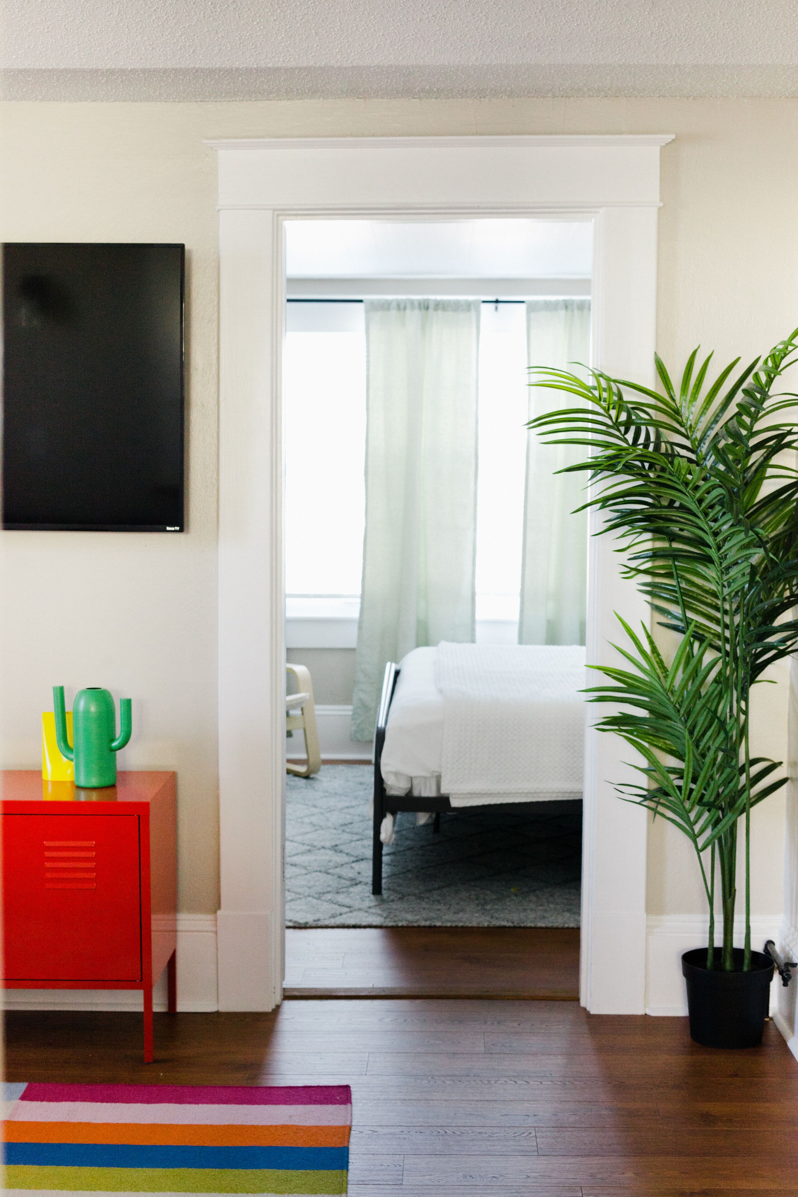 A hotel room with a bed, a potted plant, a red cabinet, and a colorful striped rug.