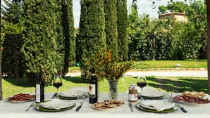 Una mesa de comedor al aire libre preparada con platos, cubiertos, vino y un jarrón de flores, rodeada de un exuberante jardín y árboles altos.