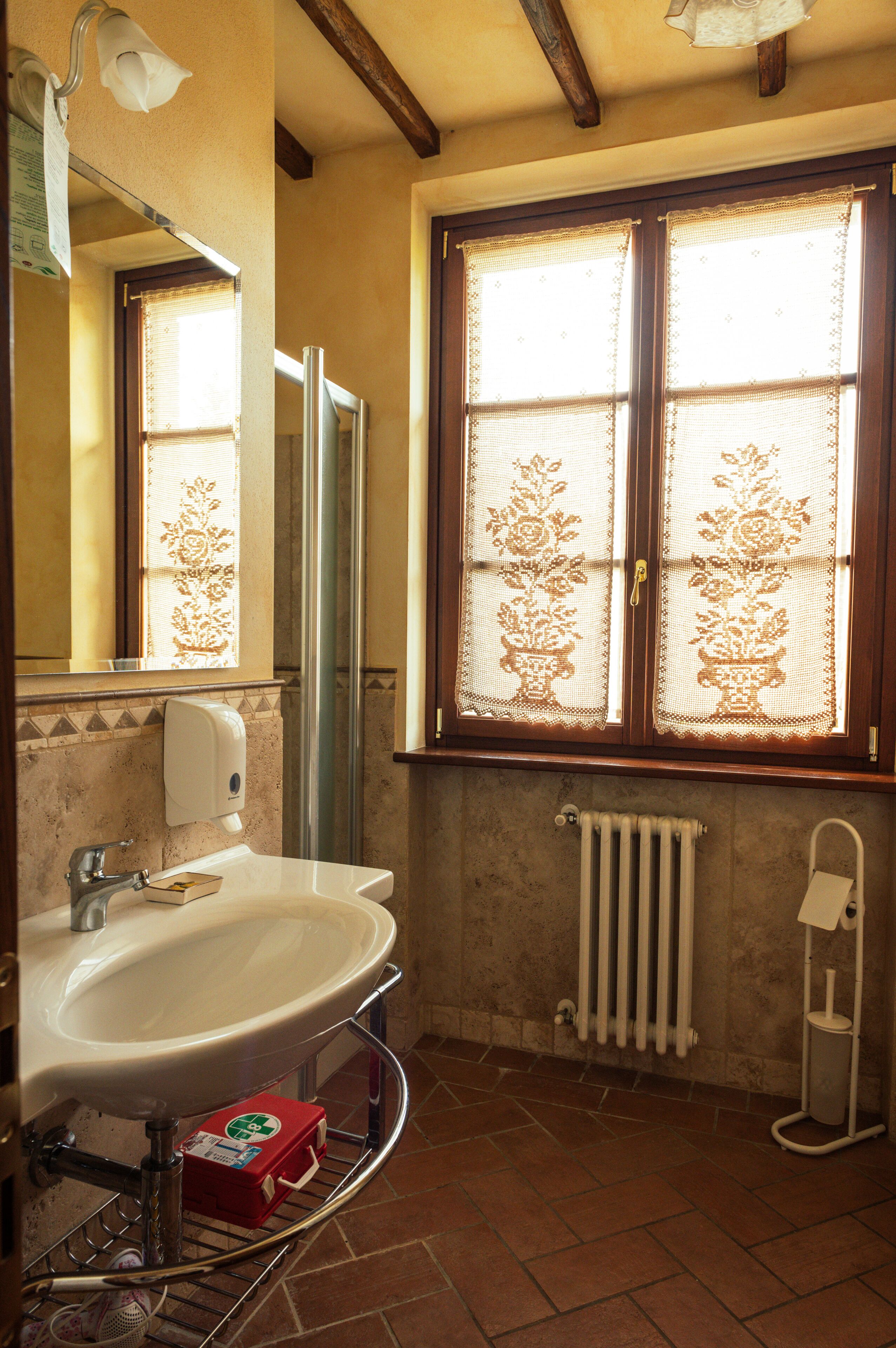 Ein Badezimmer mit weißem Waschbecken, einem Spiegel, einem Handtuchhalter, einem Fenster mit Spitzenvorhängen und einer Heizung.