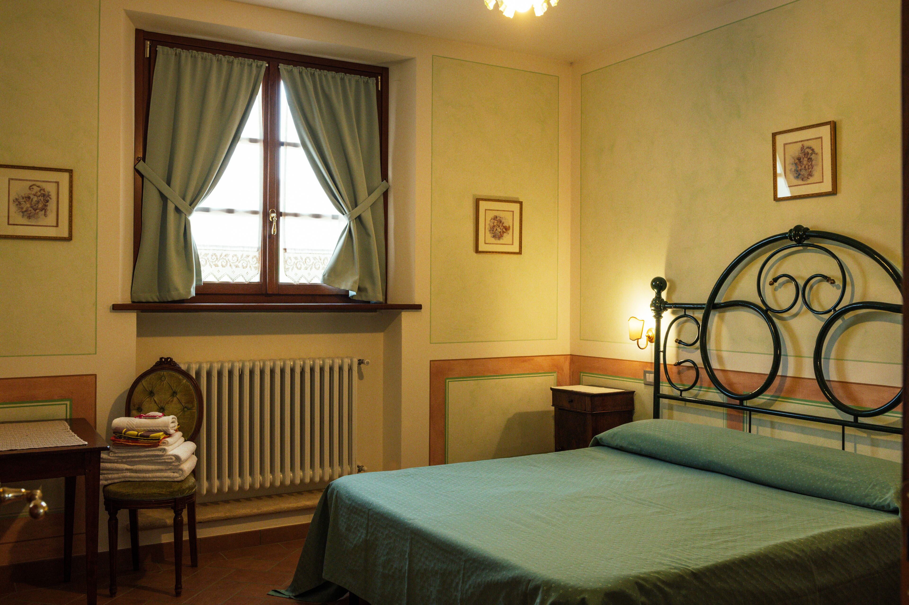 Un dormitorio con cama, una silla con toallas, un radiador, una ventana con cortinas y una pared decorada con marcos.