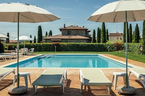 Une piscine entourée de chaises longues et de parasols, bordée d’une haie bien entretenue et avec un bâtiment en pierre en arrière-plan.