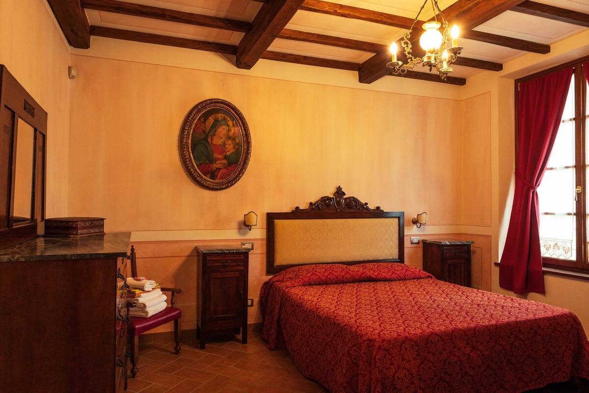 Un dormitorio con cabecera de madera, cama con colcha estampada roja, mesita de noche, cómoda, espejo y un cuadro colgado en la pared.