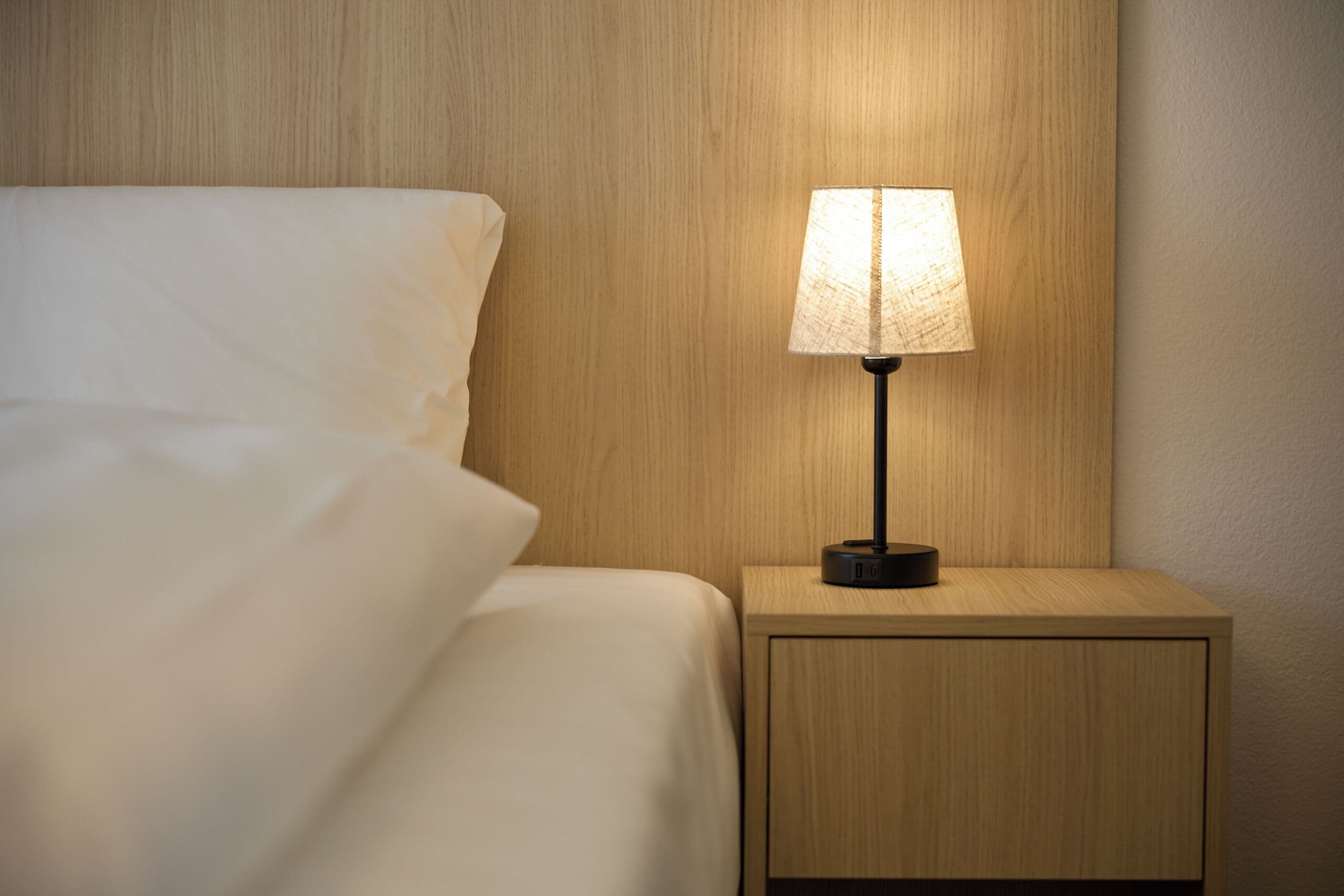 A neatly made bed with a white comforter and a wooden nightstand with a lamp.