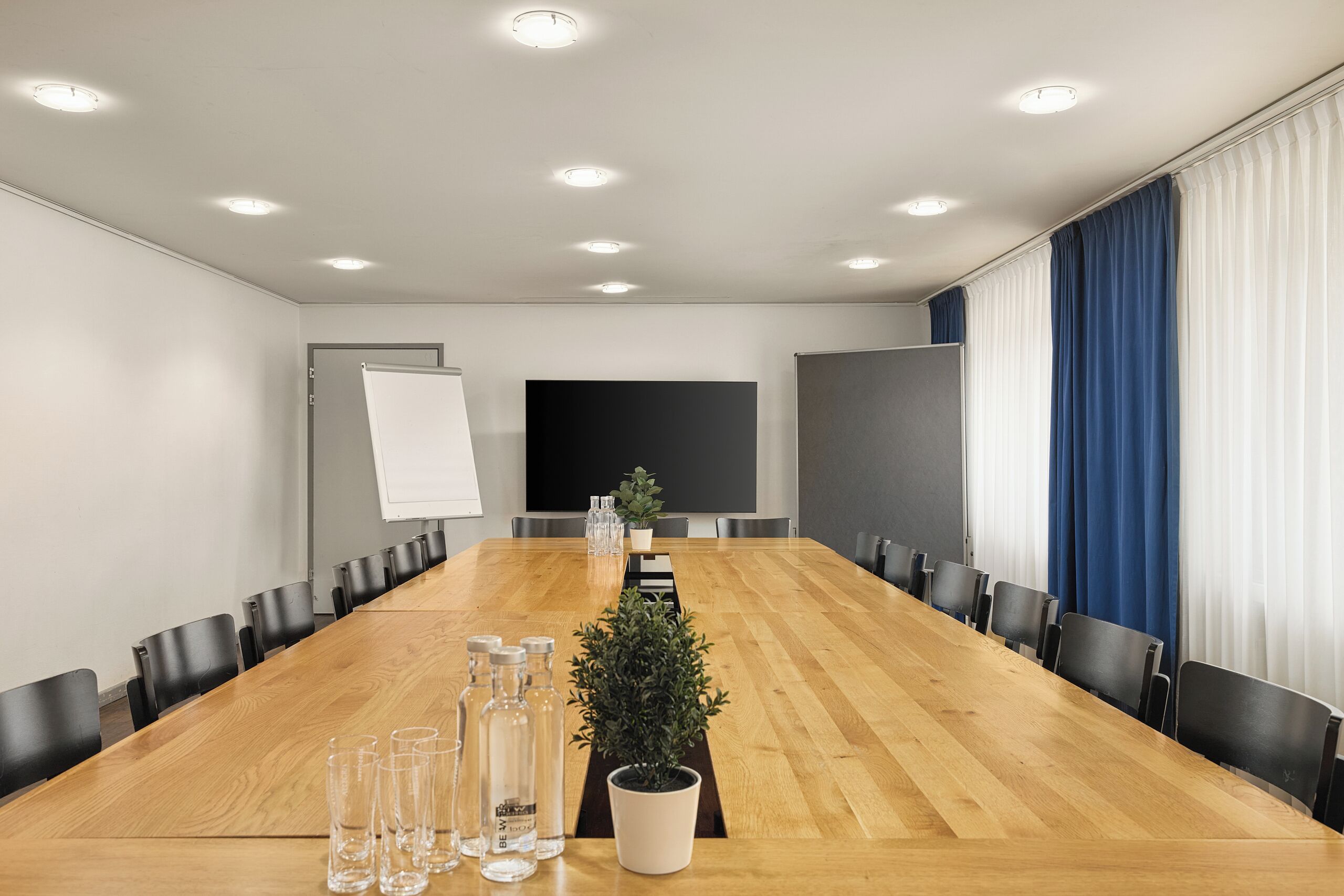 A conference room with a long wooden table, black chairs, a whiteboard, a projector screen, and a potted plant.