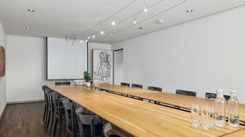 Un salón de conferencias con una larga mesa de madera, sillas negras, una pantalla de proyector y una obra de arte enmarcada en la pared.