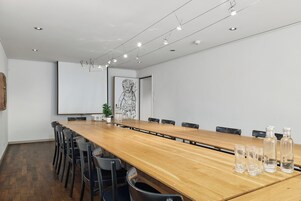 A conference room with a long wooden table, black chairs, a projector screen, and a framed artwork on the wall.
