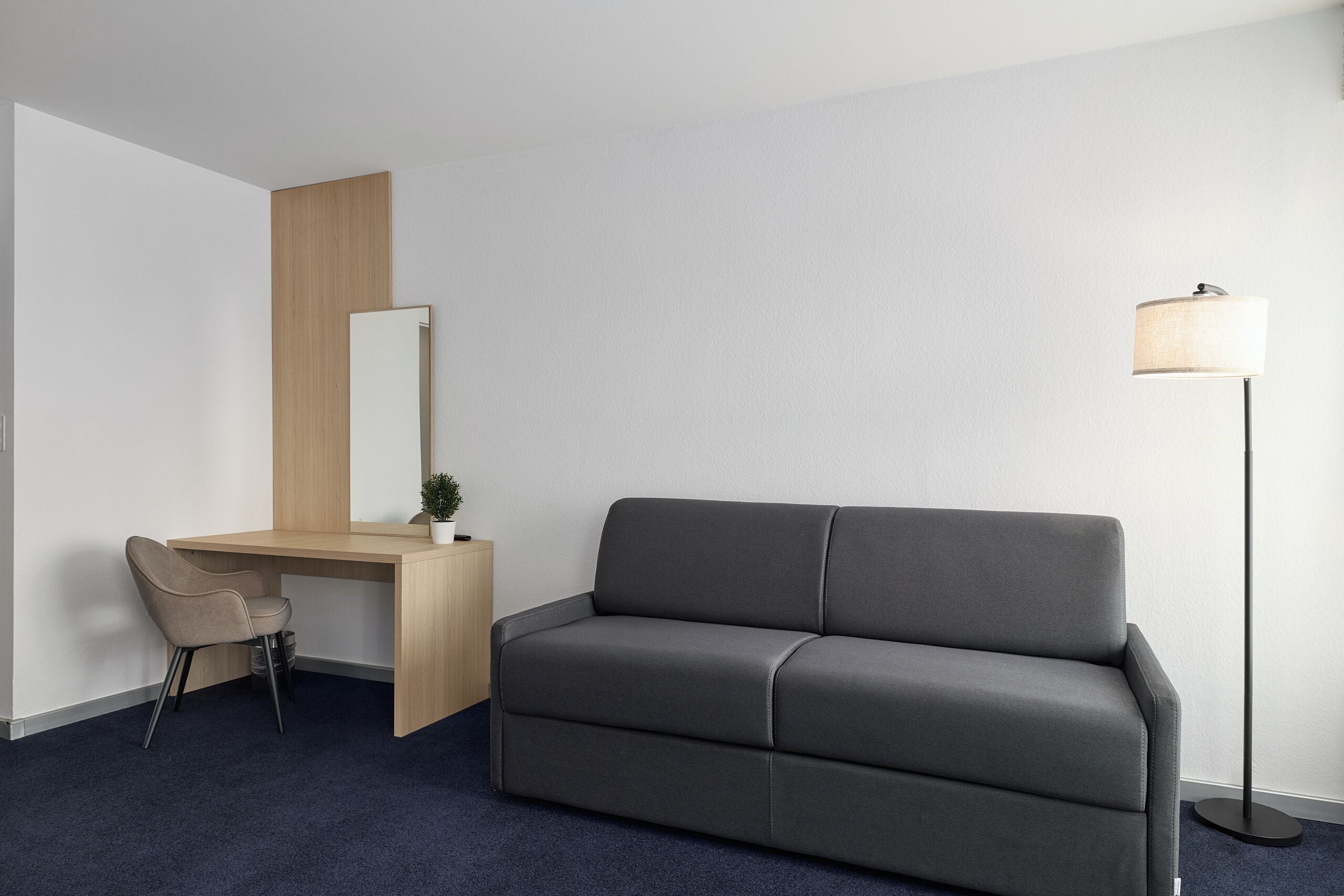 Ein modernes Hotelzimmer mit einem dunkelgrauen Sofa, einem Holztisch mit Stuhl, einer Stehlampe und einem großen Spiegel.