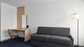 Ein modernes Hotelzimmer mit einem dunkelgrauen Sofa, einem Holztisch mit Stuhl, einer Stehlampe und einem großen Spiegel.