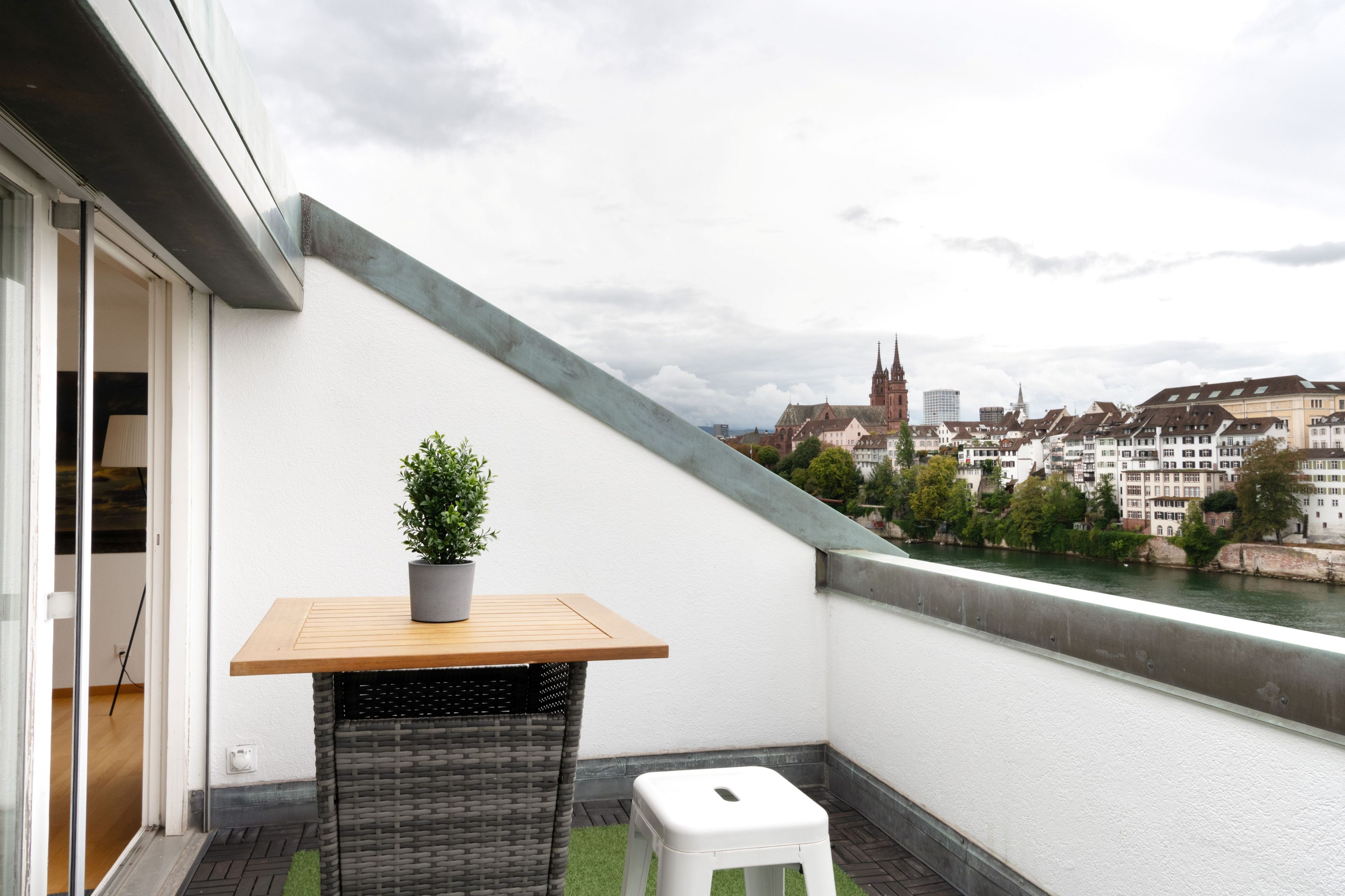 Un balcon avec une table en bois, une plante en pot et un tabouret blanc. On aperçoit une rivière et des bâtiments au loin.