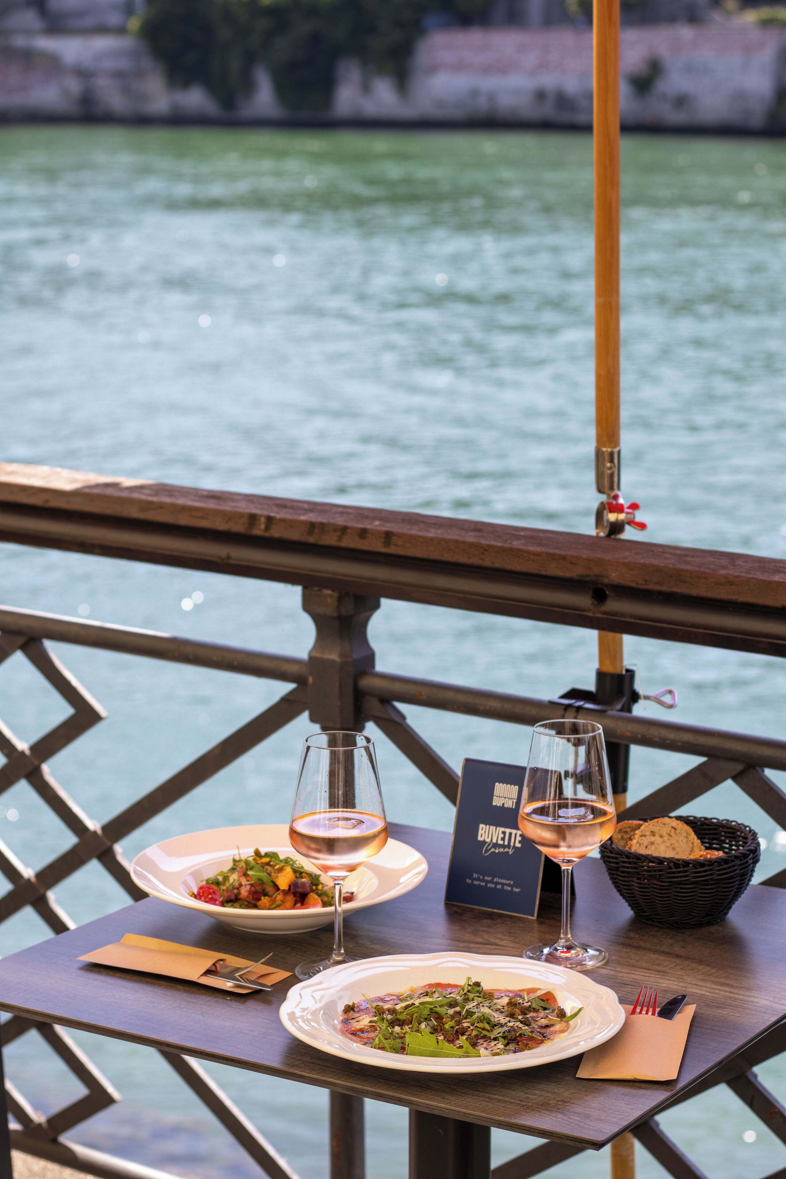 Une table avec une assiette de nourriture, un verre de vin et un menu, le tout placé sur un balcon donnant sur une étendue d’eau.