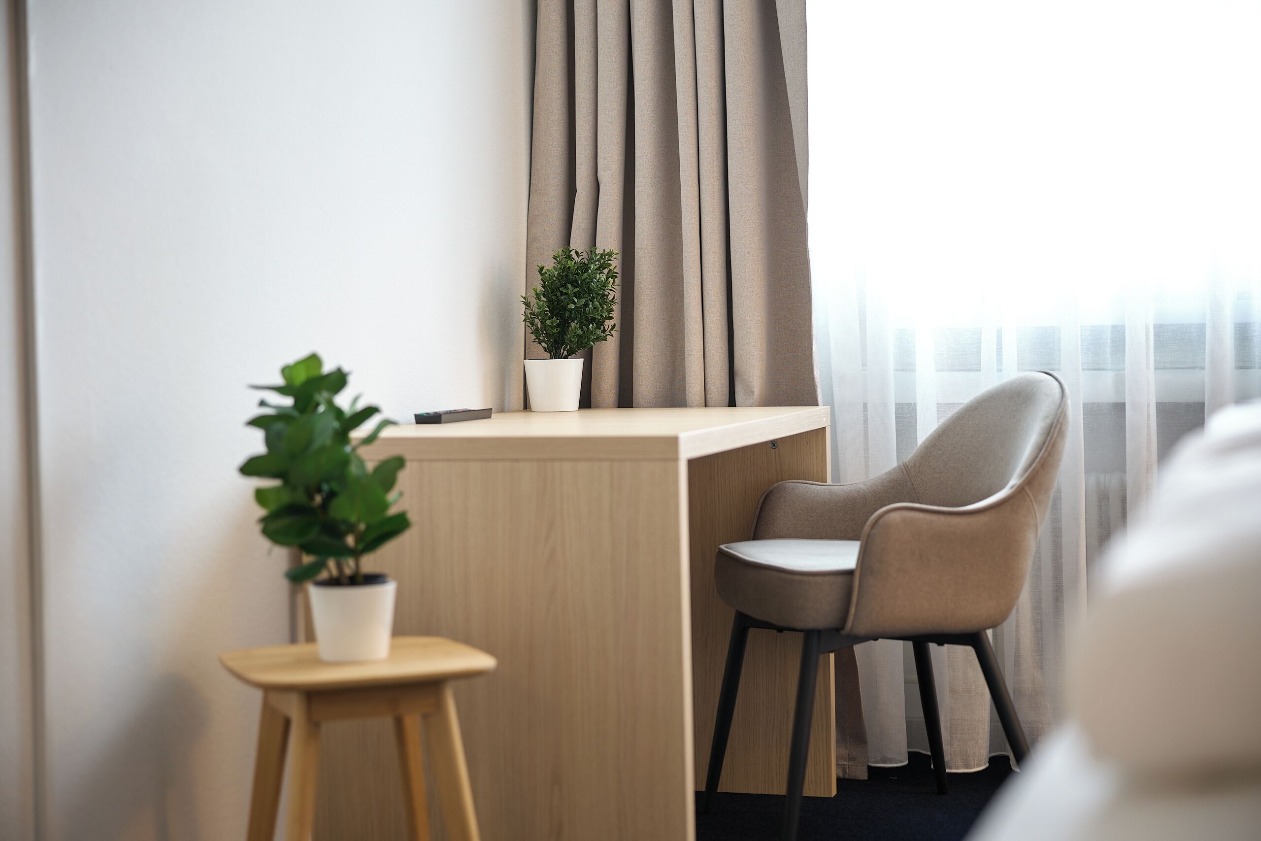 Une chambre d’hôtel moderne avec un bureau en bois, une chaise, une petite plante en pot et une vue sur la ville à travers des rideaux transparents.
