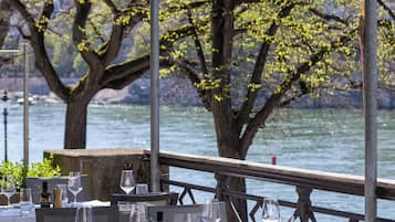 Un área de comedor al aire libre con mesas preparadas para dos, con vista a un cuerpo de agua y árboles.
