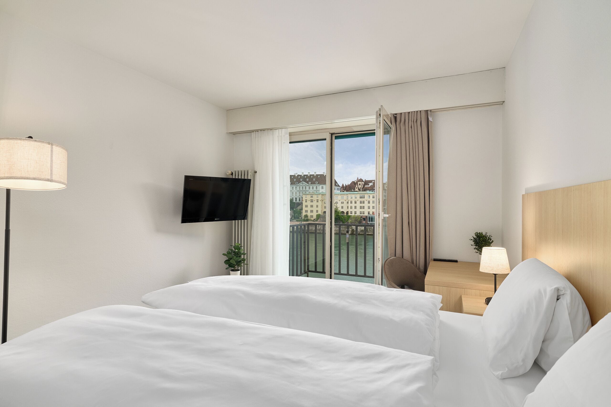 A hotel room with a bed, a TV, a desk, and a view of buildings through the window.