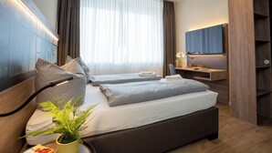 Ein modernes Hotelzimmer mit zwei Betten, einem Fernseher, einem Schreibtisch und einem großen Fenster mit Vorhängen.