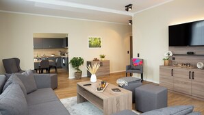 Ein modernes Wohnzimmer mit einem grauen Sofa, einem Couchtisch und einem Fernseher. Im Hintergrund ist eine Küche und ein Essbereich zu sehen.
