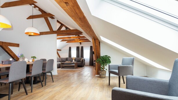 Ein geräumiges Loft mit hohen Decken, Holzbalken, einem Essbereich mit Stühlen und Tisch, einer Sitzecke mit Sofa und einem großen Fenster für natürliches Licht.