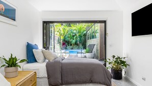 A bedroom with a bed, a small table, and a chair. There is a sliding glass door leading to a balcony with a pool and greenery.