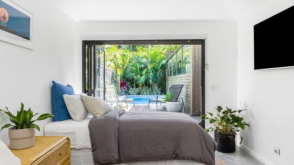 A bedroom with a bed, a small table, and a chair. There is a sliding glass door leading to a balcony with a pool and greenery.