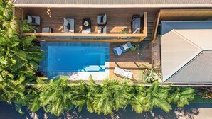 An aerial view of a modern outdoor pool area with lounge chairs, a wooden deck, and lush tropical plants.