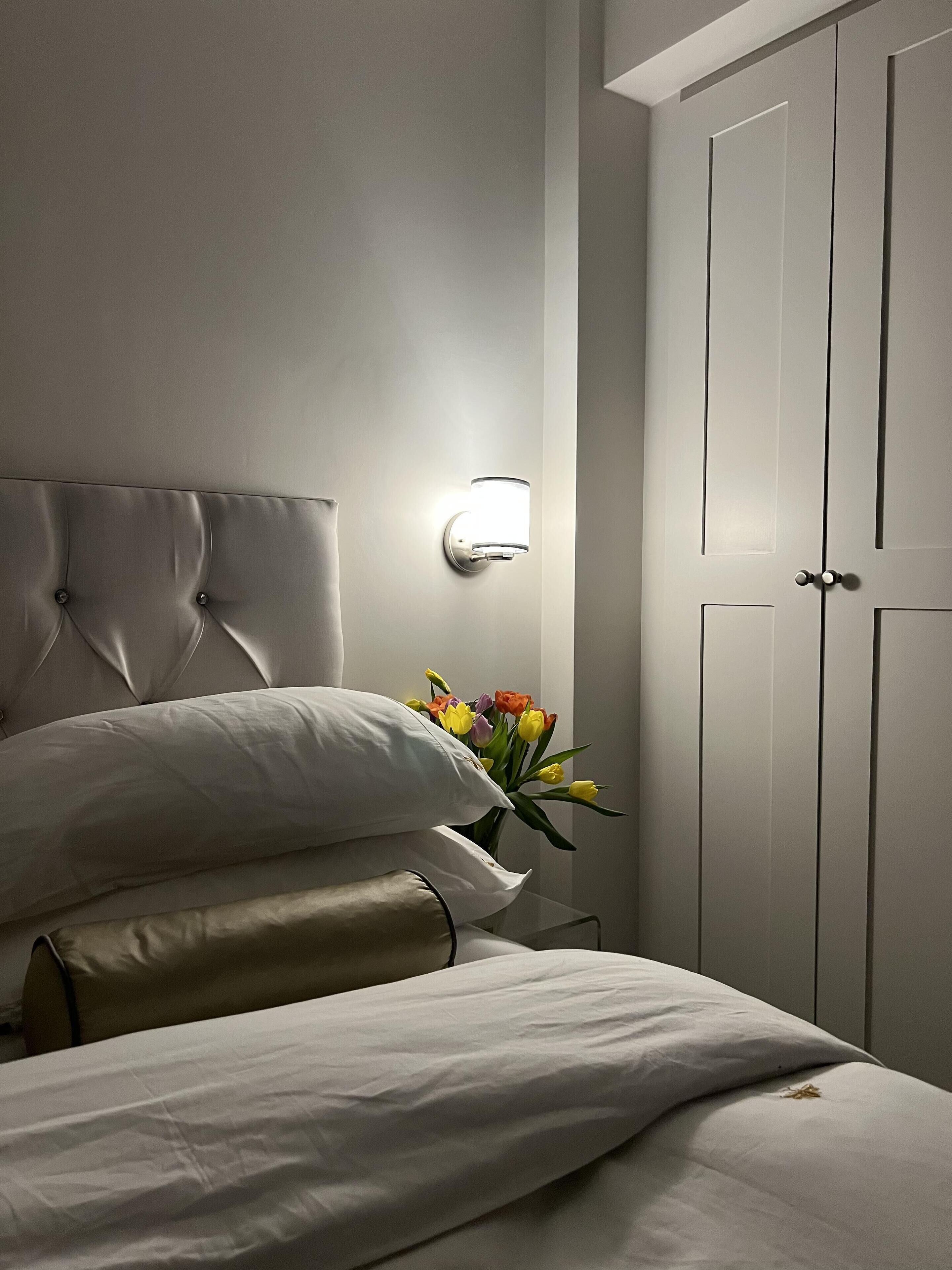A neatly made bed with white linens, a wall-mounted light fixture, and a vase with flowers.