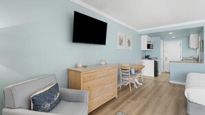 Un salon comprenant un canapé, un téléviseur fixé au mur, une commode en bois et un coin repas avec des chaises et une table.