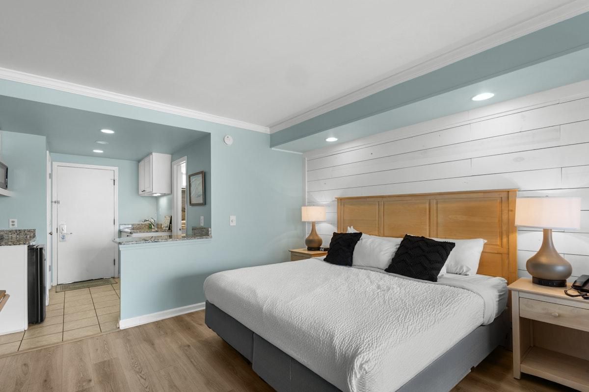 Une chambre avec un grand lit, des tables de chevet, une tête de lit en bois et, au fond, un coin cuisine.