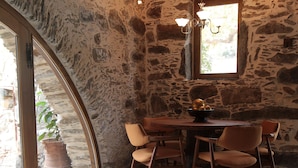 Un interior rústico de piedra con una mesa y sillas de madera, una vela sobre un tocón y una ventana con luz natural.