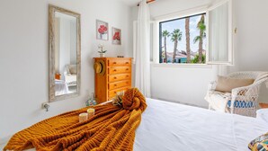 A bedroom with a bed, a dresser, a window with a view of palm trees, and a wicker chair.