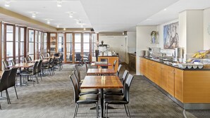 A spacious dining area with multiple tables and chairs, a kitchen counter with food items, and large windows allowing natural light. - Panoramic Perth Views: 1BR on Mounts Bay Road (Perth)