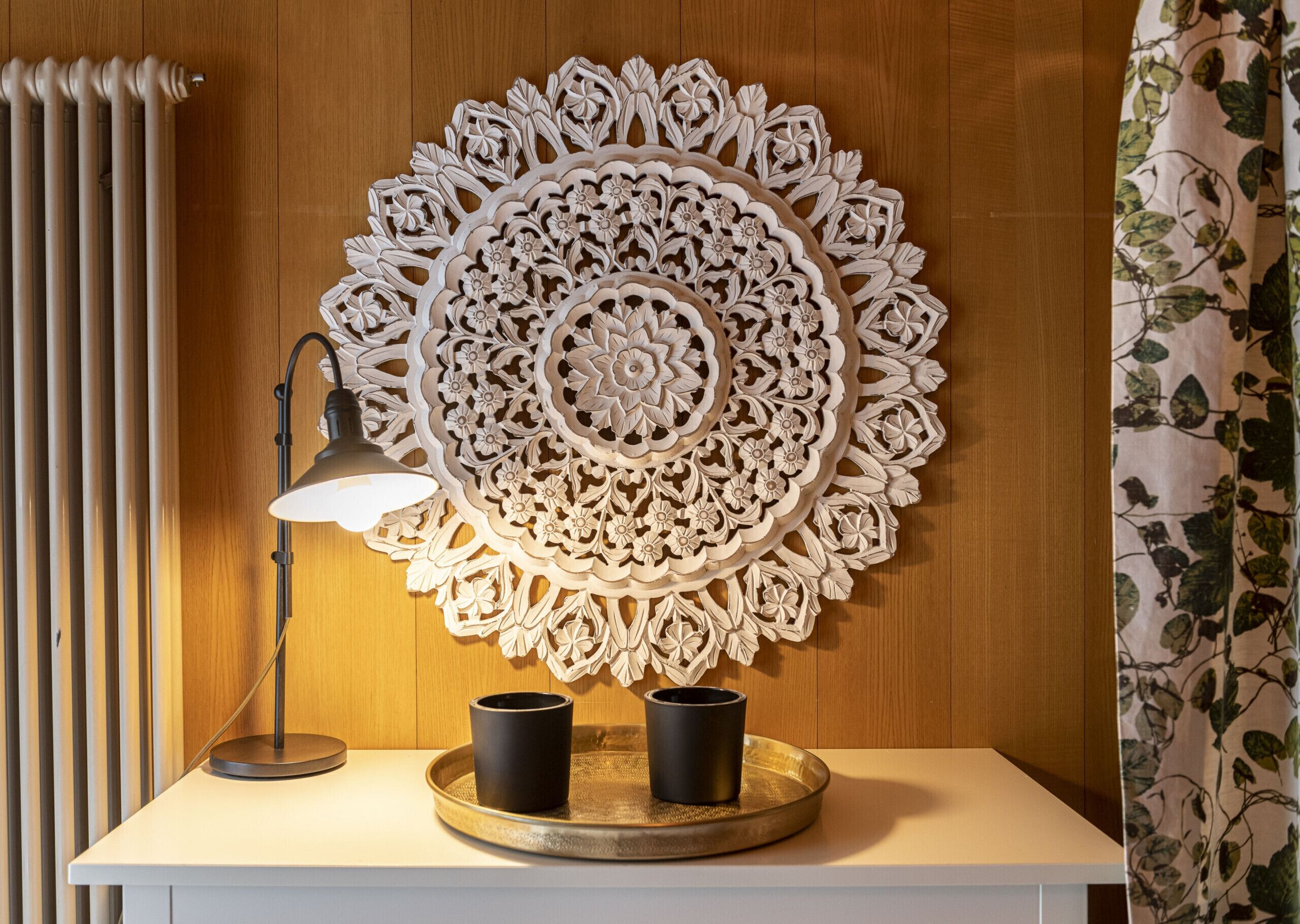 A wall-mounted decorative piece with a circular pattern, a desk lamp, two black cups on a tray, and a radiator.