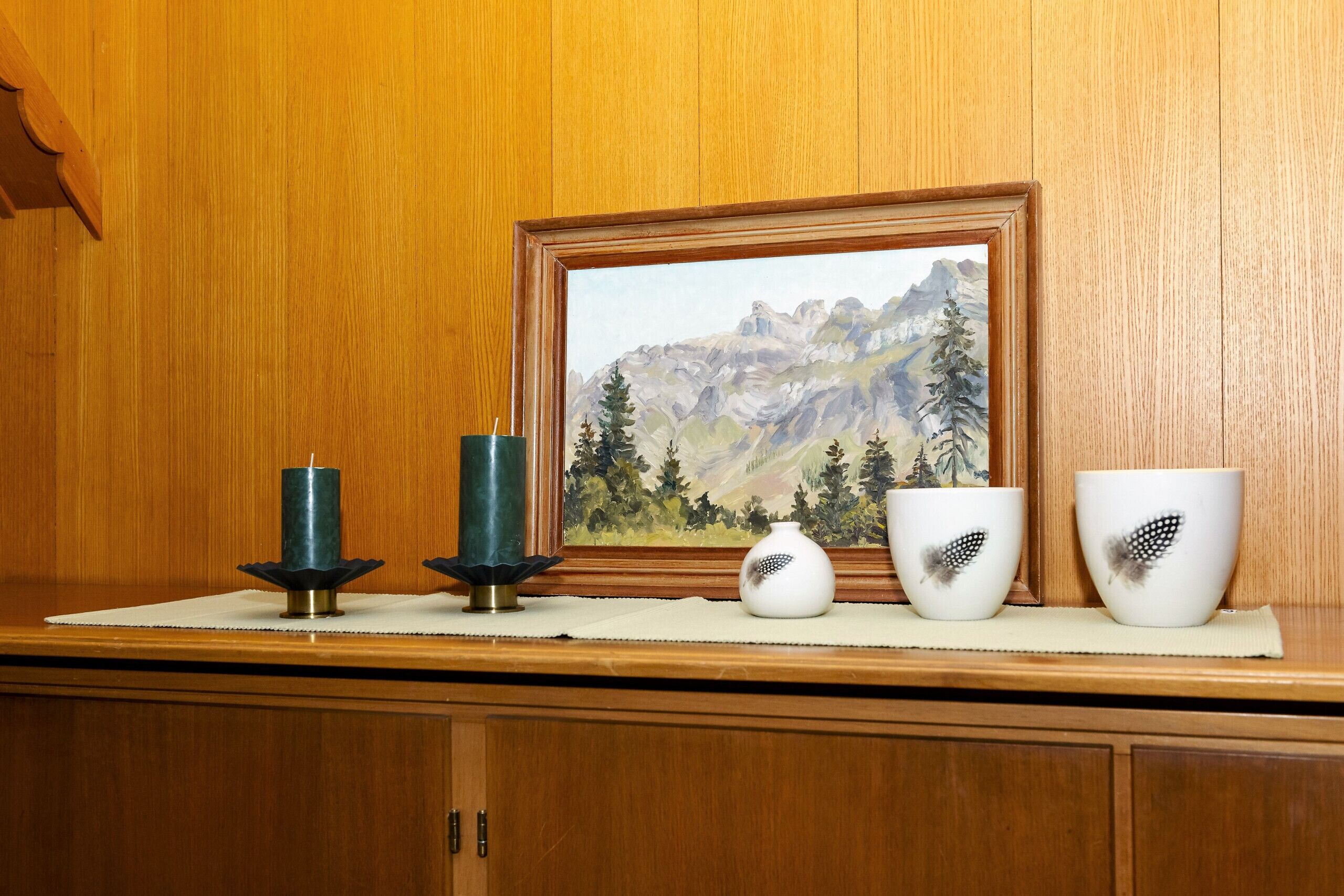 Um armário de madeira com um quadro emoldurado de uma paisagem montanhosa, duas velas e três tigelas decorativas sobre um jogo americano.