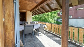 Un balcón exterior con techo de madera, una mesa con sillas y vistas a zonas verdes y edificios al fondo.