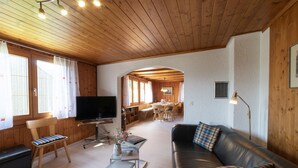 Una sala de estar acogedora con un sofá de cuero, un televisor, una zona de comedor de madera y una cocina al fondo.